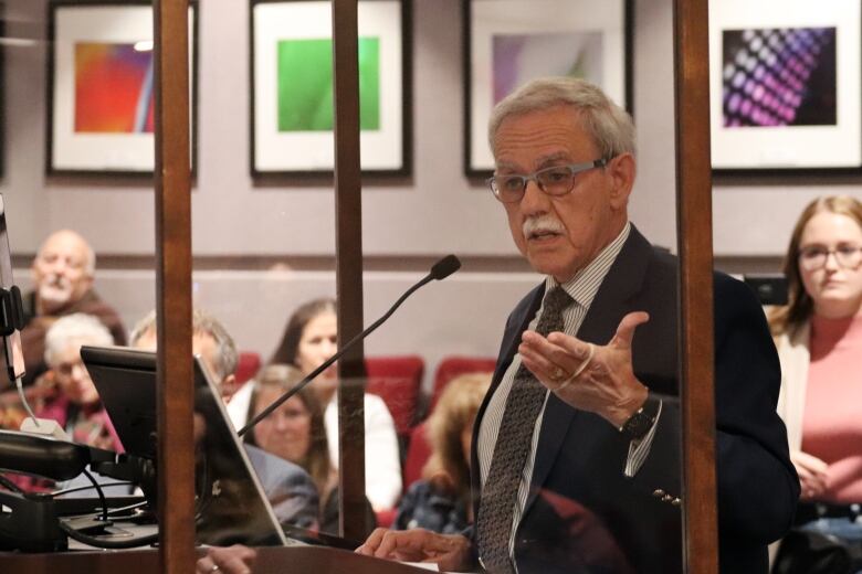 A man with silver hair and mustache addresses a city council chamber, with people sitting in the background.