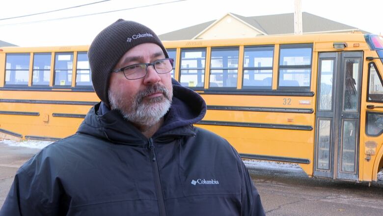 Middle-aged man with a beard and glasses wears a black hat and jacket and stands infront of a yellow school bus. 