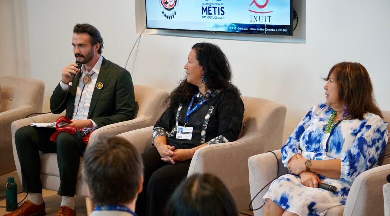 Three people sit in chairs in front of a crowd.