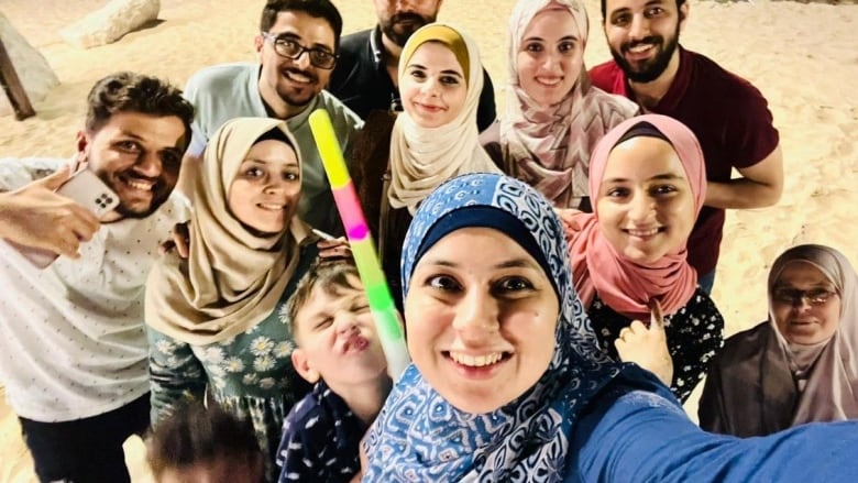 A smiling woman in a blue headscarf snaps a selfie with two little boys beside her making funny faces, and nine other smiling family members.