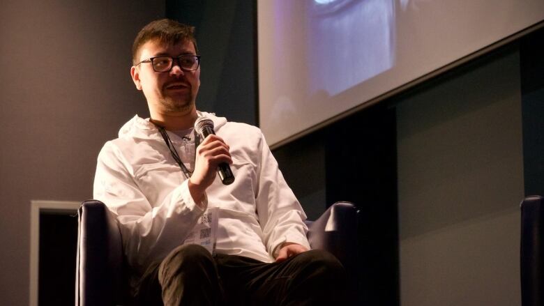 A young Inuit man wearing a white zip-up jacket and a lanyard with a conference badge sits in an armchair on a stage and speaks into a microphone