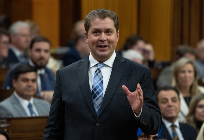 A man in a dark suit and blue tie rises in the House of Commons.