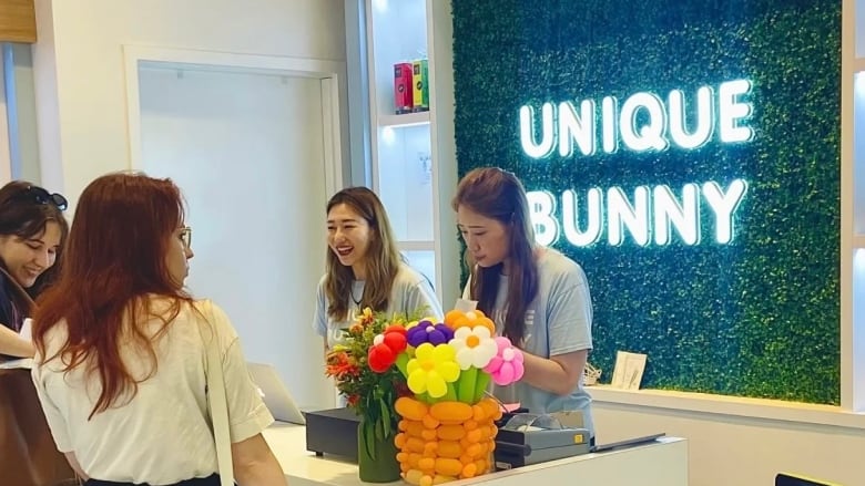 Two women are pictured standing behind the cash register at a store as customers stand on the other side of them. A sign that says 'UNIQUE BUNNY' in LED lights is seen behind them.