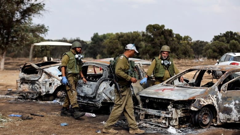 Three soldiers in green uniforms and helmets walk around the remains of burnt cars.