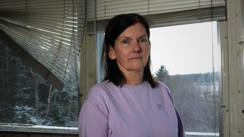 A dark-haired woman in a mauve sweater stands in the livingroom of a dilapitated mini-home. 