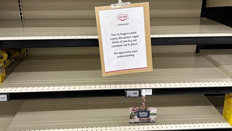 Empty shelves at a grocery store can be seen there is a sign hanging on one of the shelves. 