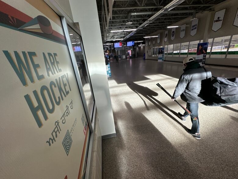 A person carrying a hockey bag and stick walks past a sign advertising We Are Hockey.