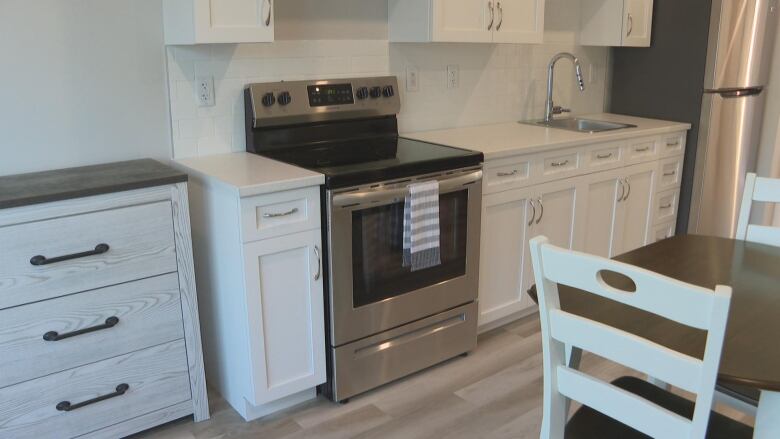 A new kitchen with a small kitchen table. The cupboards are white. The oven is silver. There is a silver sink and light brown floors. 