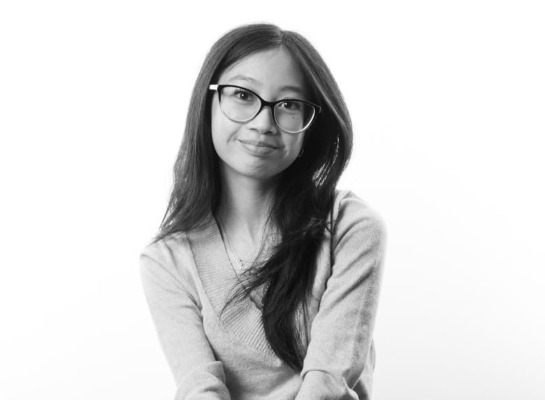 Black and white portrait of young woman with long, straight dark hair and round glasses. She is wearing a V-neck top and jeans and sits on a stool. Her legs are crossed and she has her hands crossed on her knee. 