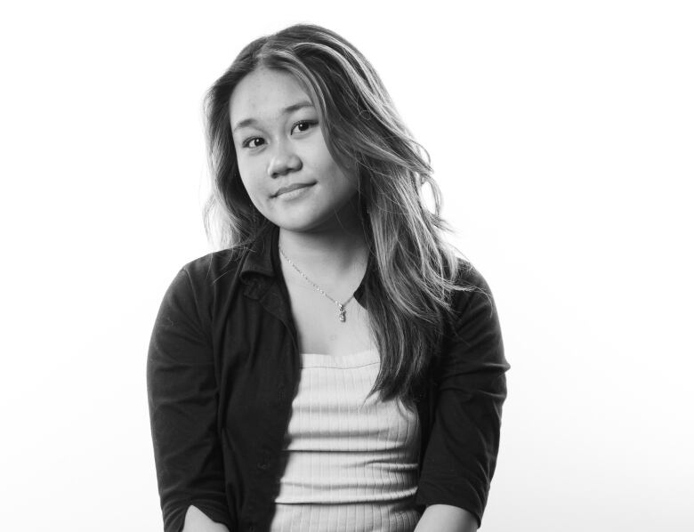 Black and White portrait of young woman with long dark hair with lighter streaks. She sits on a stool and is wearing a light-coloured tank top underneath a button-down, collared top. She wears dark cargo pants and has her hands clasped on her lap. 