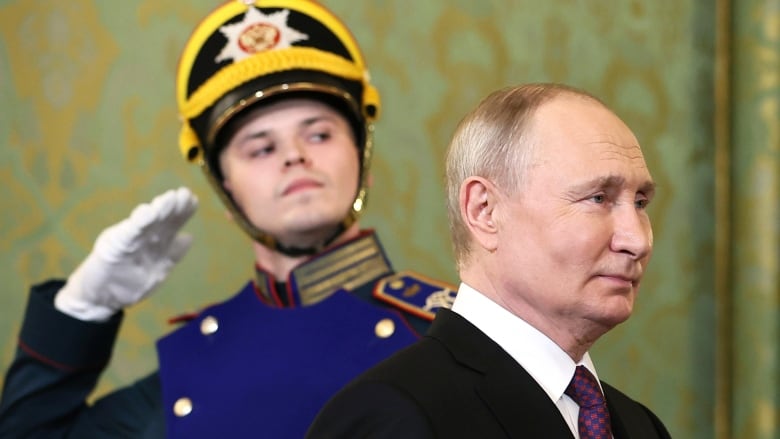 An older, clean shaven man with a receding hairline wearing a suit and tie, is shown in profile. Behind him, is a military guard wearing a helmet.