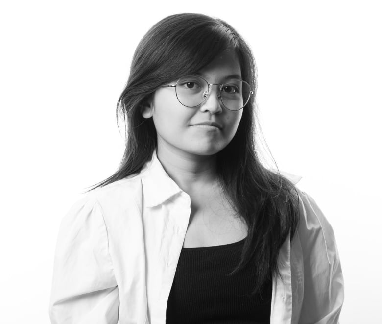 Black and white portrait of a young woman with long, straight dark hair. She has round glasses and is wearing a black tank top underneath a light-coloured button down shirt. She is wearing light-coloured pants and bracelets around her right wrist. 