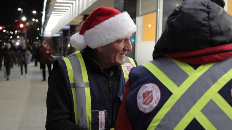 a man in a santa hat and bright vest smiles on a busy sidewalk. 