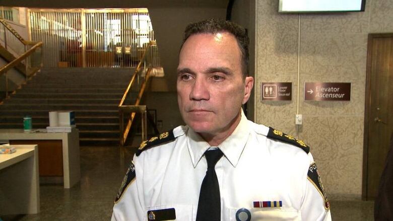 A man with short dark hair, wearing a white shirt with patches on the shoulders, stands in the lobby of a building.