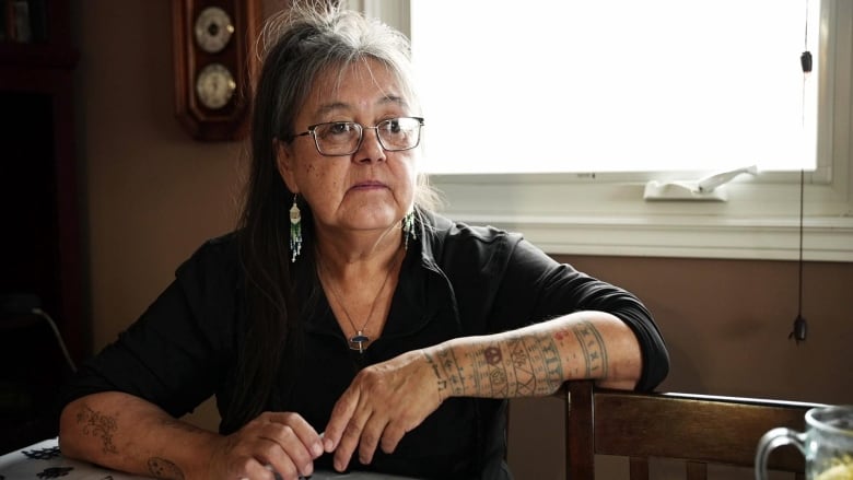 A woman sits at a table with a window behind her. She has a variety of traditional Indigenous tattoos visible on her arms. 