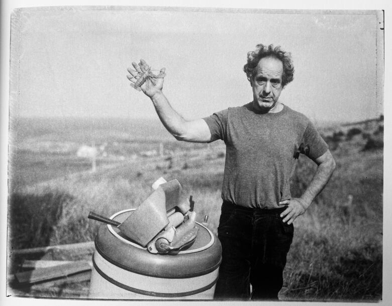 In this black-and-white image, a man wearing a t-shirt stands outside next to a stand-up washing machine. He extends the palm of his right hand towards the camera.