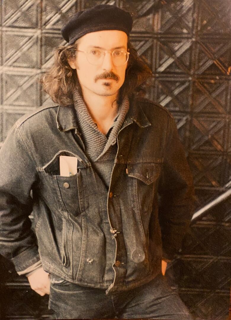 A man wearing glasses with long curly hair poses for a portrait in a jean jacket with black beret on his head. He stands in front of a wall of glass bricks, with an envelope sticking out of his breast pocket.