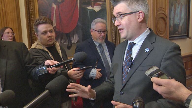 A man in a suit surrounded by reporters with microphones and tape recorders pointed toward him speaking with his hands raised.