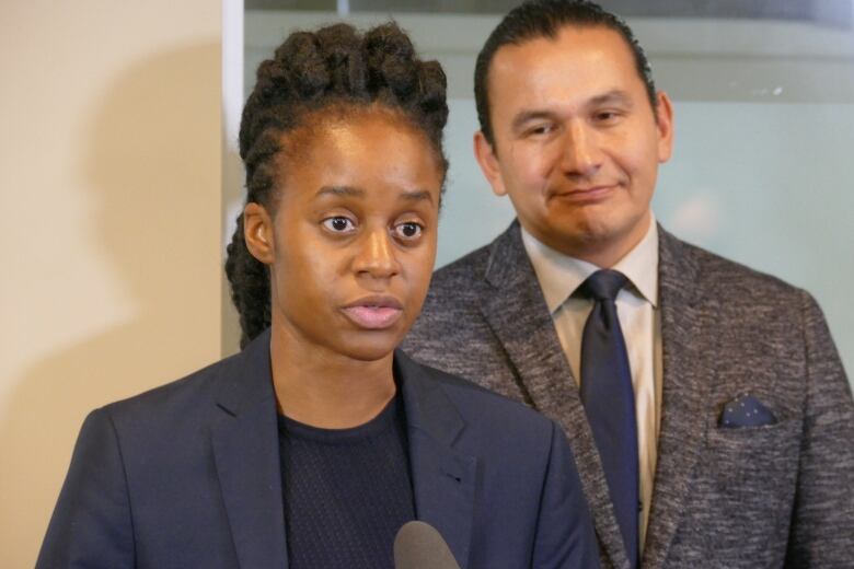 Two people stand at a podium. The one closest to it has long black hair in braids. The one in the background wears a suit and had black hair pulled back in a braid.