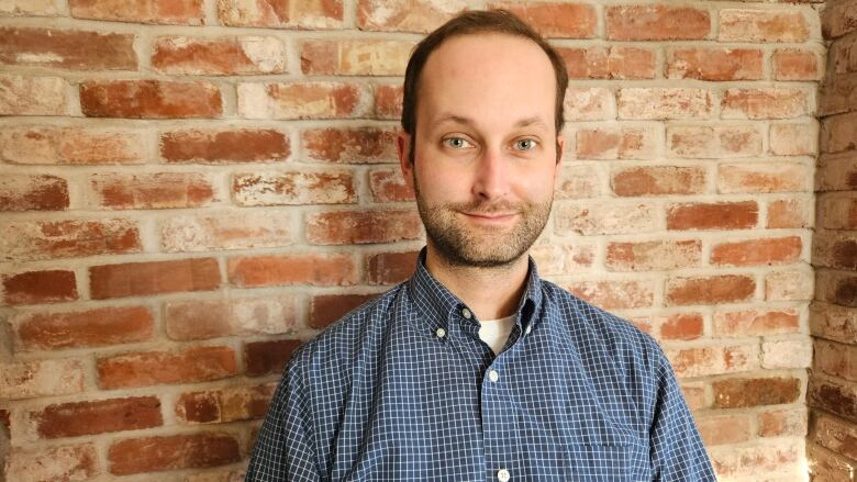 Head and shoulders shot of a man with a beard and mustache in a checked shirt, against a brick wall.