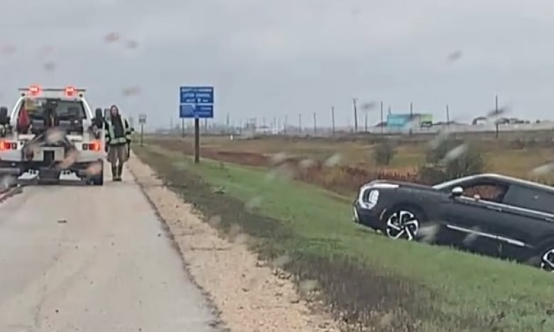 A black SUV lies in a green grassy ditch with the hood pointing upward toward the highway. The sky is grey. On the left of the photo, which was taken through a windshield with raindrops on it, is a tow truck with a tow truck driver standing beside it.