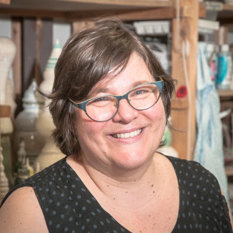 A white woman with glasses and short brown hair wears a black polkadot shirt. Wooden shelves and art supplies are behind her