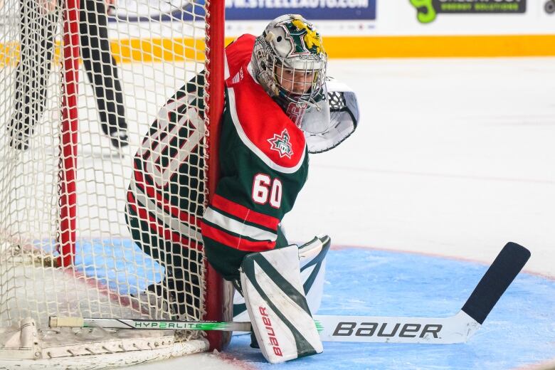 A junior hockey goalie wearing a red and green jersey is shown looking to the side of the net.