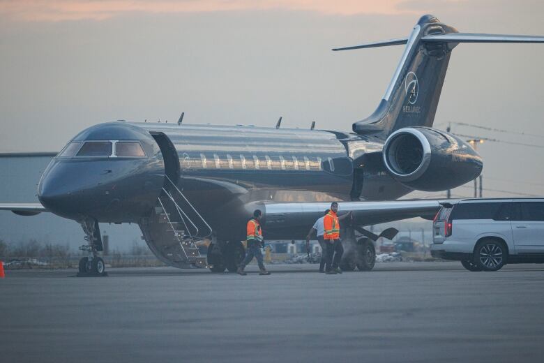 A private jet with its staircase descended as two airport workers walk next to it.