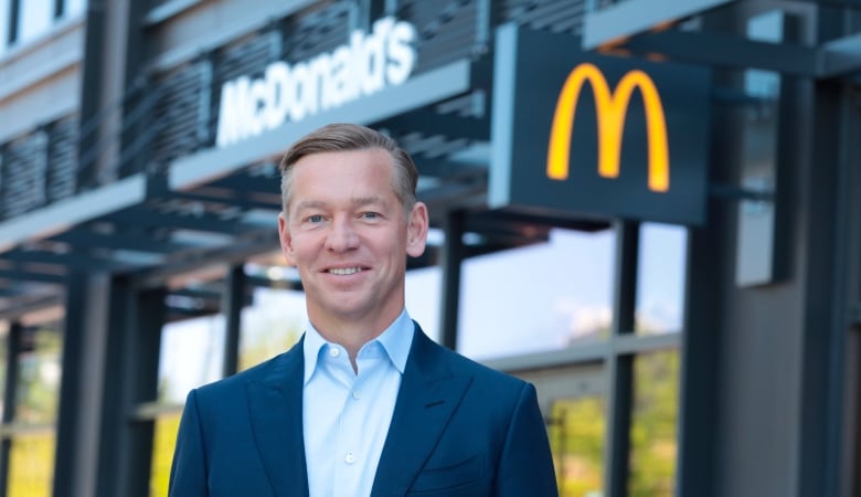 A man with short brownish-grey hair wears a blue shirt and a navy blazer as he stands in front of a McDonald's restaurant.