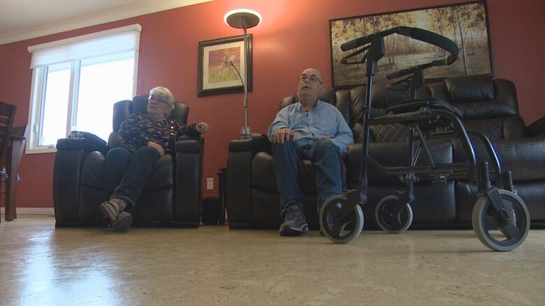 A woman and a man sit on a couch, while a walker is stationed in the foreground. 