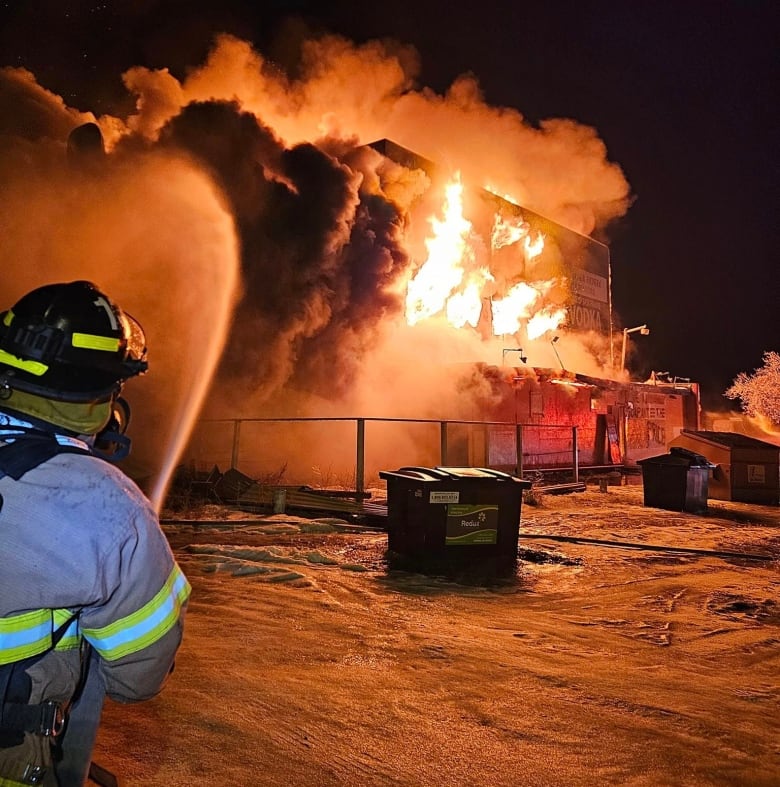 it's night and a firefighter in full gear is holding a hose with water blasting outwards toward a building on fire