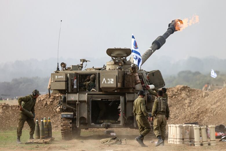 A tank fires a round as soldiers load munitions and watch on.