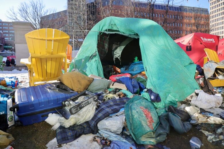 A camping tent covered in a green tarp sits on the ground in Halifax's Grand Parade square. The entrance to the tent is open and inside is charred black from a fire. Dozens of items are strewn across the floor of the tent and spill out onto the ground around it.