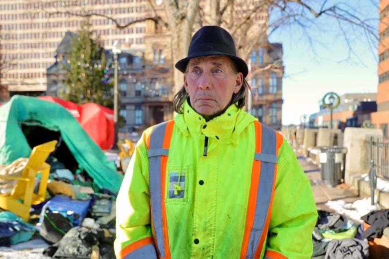 A man in a black fedora and neon jacket stands in front of a burned out tent at Halifax's Grand Parade.