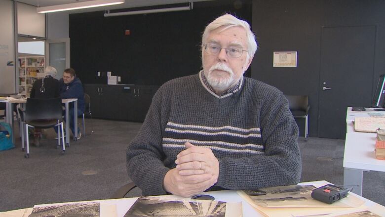 A man in a grey sweater speaks to the camera with a table covered with old photos in front of him.