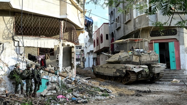 Three soldiers stand beside a severely damaged building. To their right is a tank, moving along a narrow street, that is filled with damaged buildings and debris.