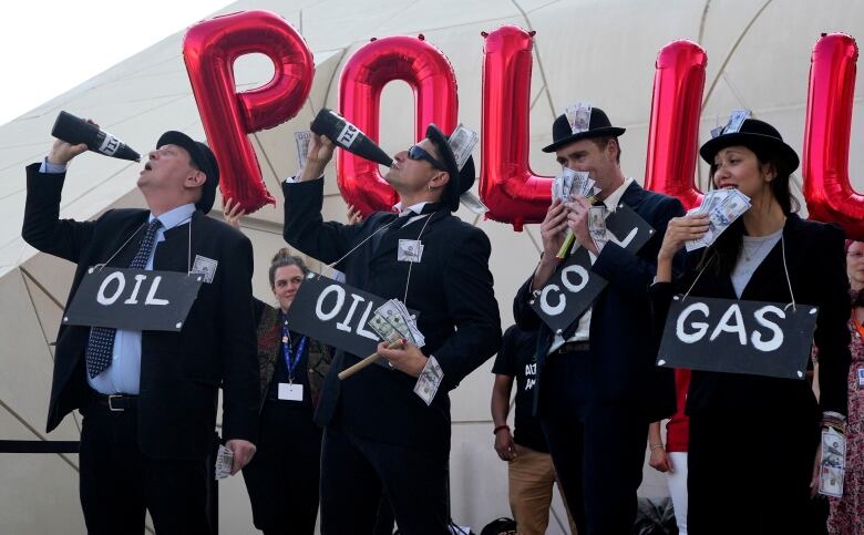 Demonstrators wear black suits and pretend to drink oil from black bottles.