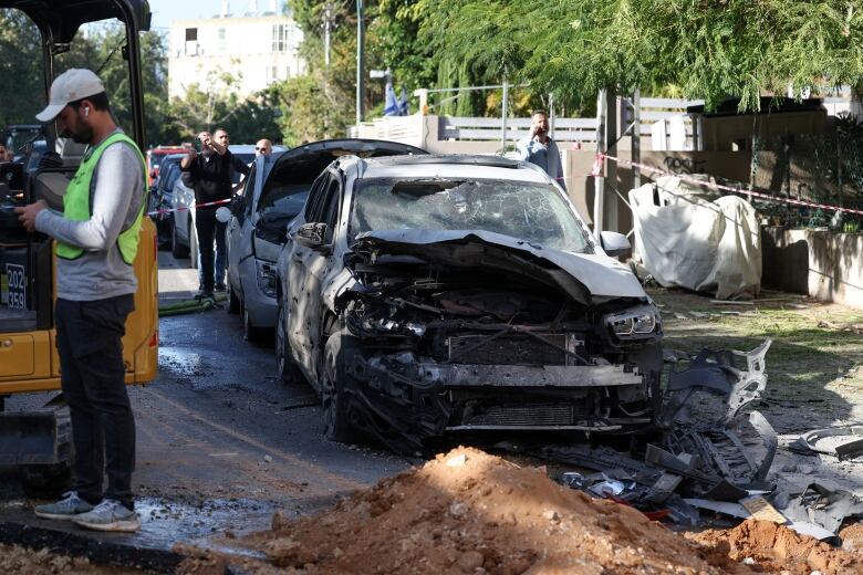 A man in a reflective vest and ballcap stands near a heavily damaged car.