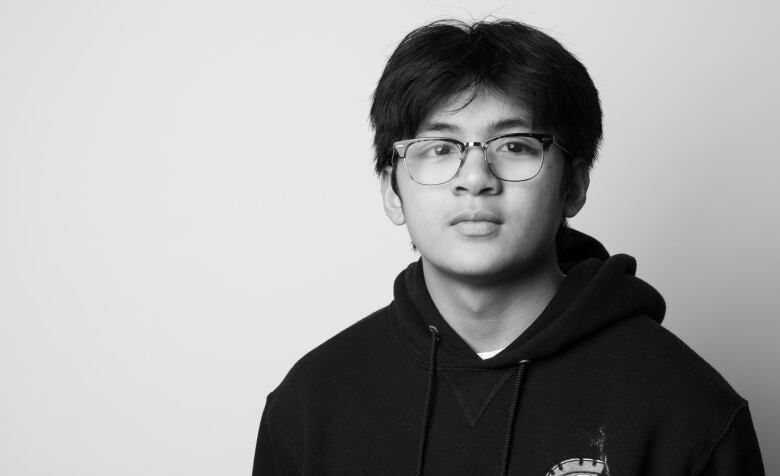 Black and white portrait of young man. Head and shoulders only portrait. Young man has dark,  shaggy hair with horn-rimmed glasses. He wears a dark sweatshirt. 