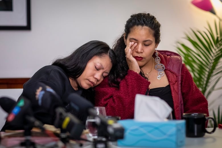 Two Indigenous women console each other as they sit in front of microphones at a press conference.