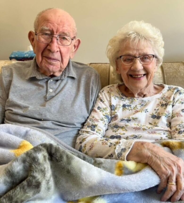 An elderly man and woman both smile as they are pictured sitting next to each other on a couch with a blanket partially covering them.