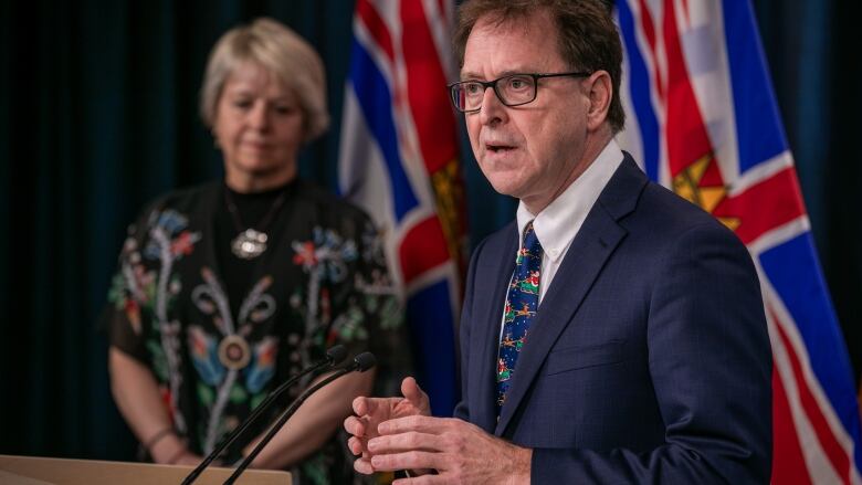 A middle-aged white woman with a blonde bob hair cut wears a black skirt and black top with a floral pattern while standing to the right of a man who is speaking from a podium. He is a middle-aged white male wearing glasses, a navy blue suit and a tie with images of Santa on a sleigh on it.