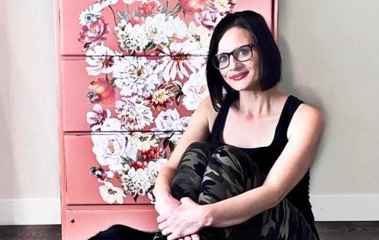 A woman sits in front of a floral painted dresser