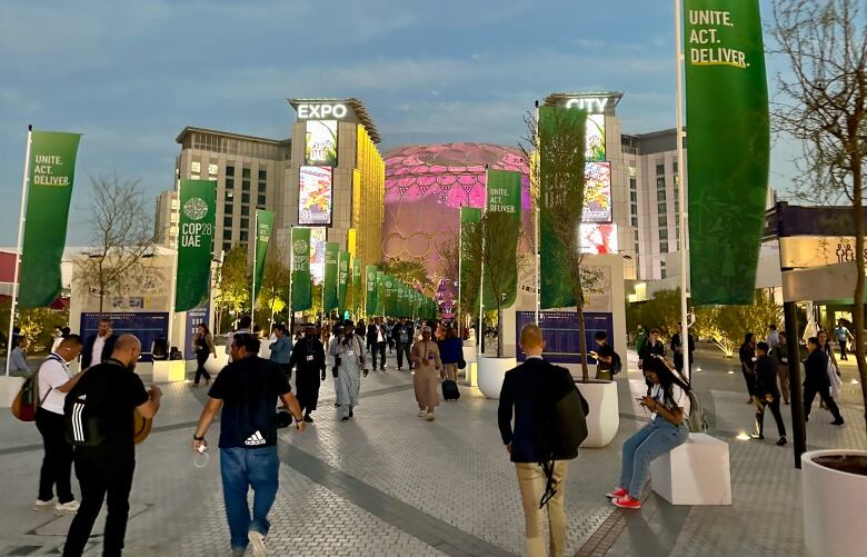 A large crowd of people walk outside through a plaza with green banners hanging above.