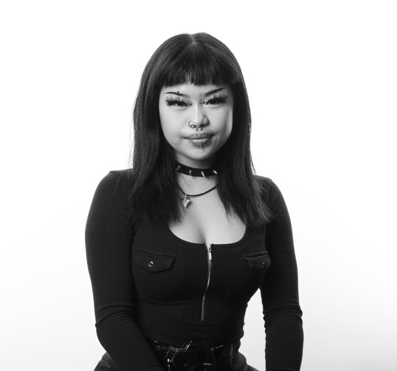 Young woman with straight, dark hair sits on stool. She has multiple face piercings including her nose and both eye brows. She wears a silver spiked collar around her neck. She is wearing a scoop neck dress with a zipper down the front. She is wearing fish net stockings. 