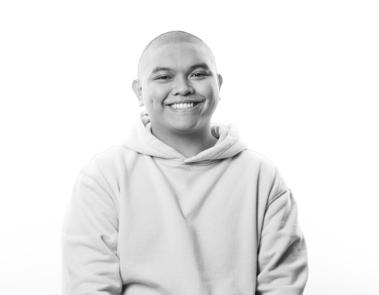 Black and white portrait of young man sitting on a stool. He is smiling broadly and has a shaved head. He is wearing a light-coloured hooded sweatshirt. 