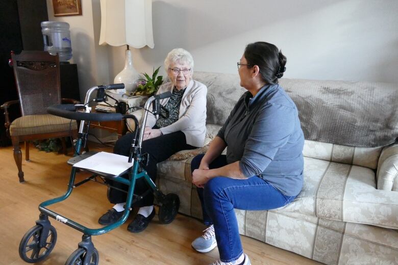 An elderly woman with a walker sits on a couch left of her home care worker in the living room of an apartment building suite.