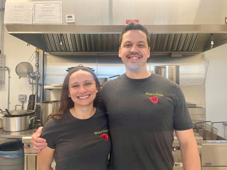 Rachel Ross and Richard Charron stand smiling in the kitchen with black shirts that say 