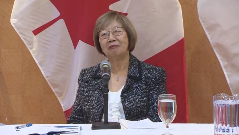 A woman wearing a tweed blazer with a white top sits at a table behind a microphone,