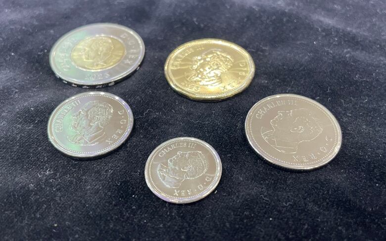 A set of five coins, each with a portrait of King Charles, is laid out on a dark service.